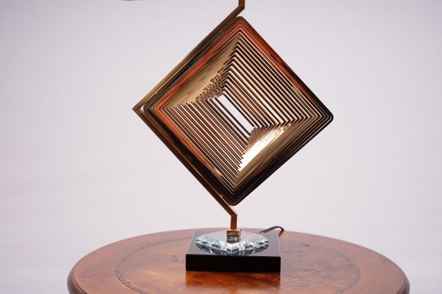A modern table lamp with a unique geometric design, featuring a gold metal base in the shape of a rotated square with intricate latticework, complemented by a sleek black glass base. The lamp is placed on a round wooden table.