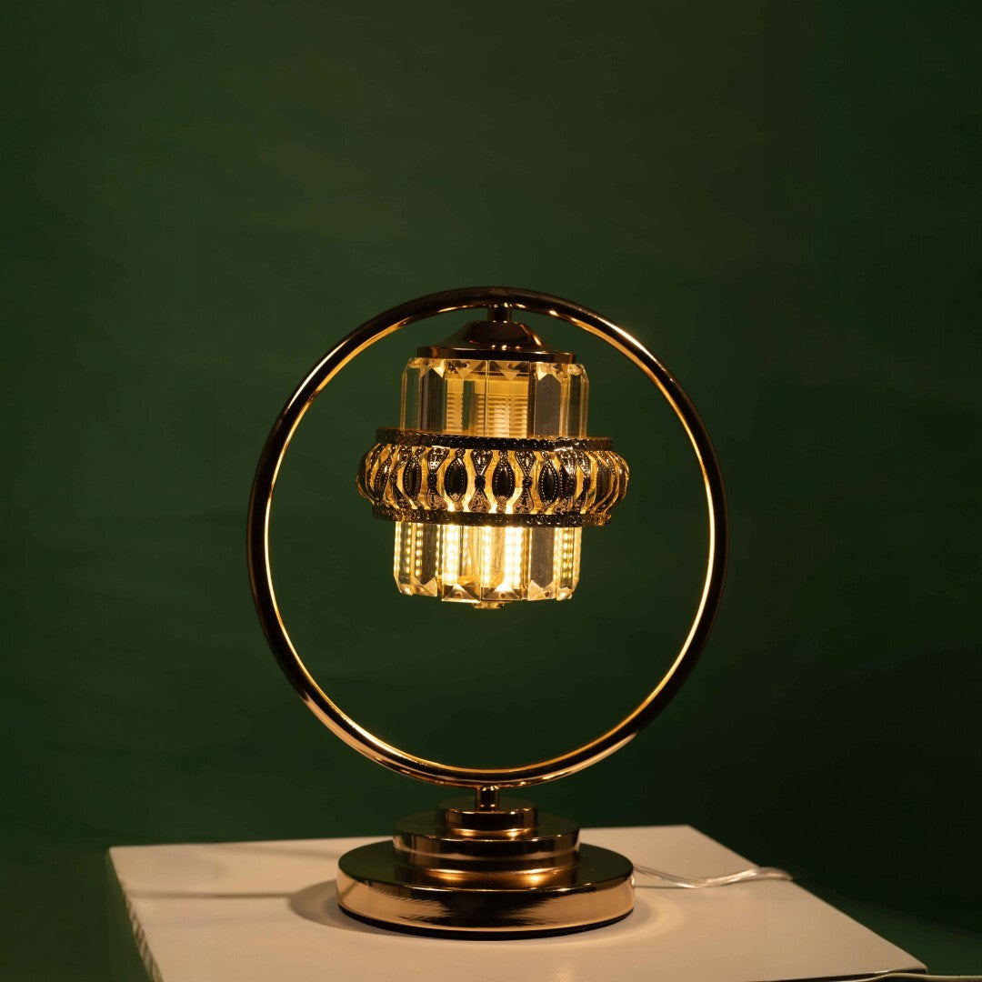 A luxurious table lamp with a circular gold frame and suspended crystal elements, equipped with led lamp E27 designed to resemble a chandelier. Placed on a white table and green background.