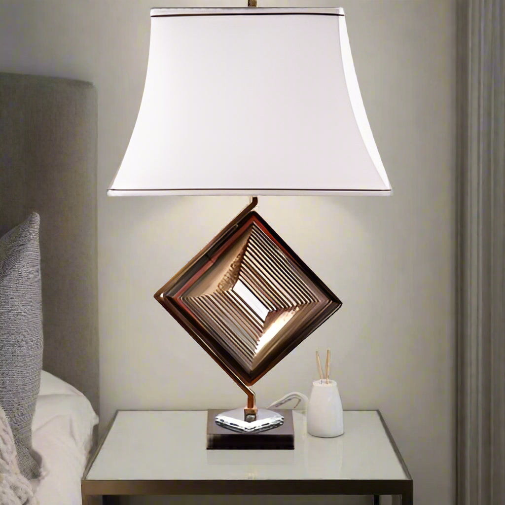 A modern table lamp with a unique geometric design, featuring a gold metal base in the shape of a rotated square with intricate latticework. The lamp is placed on a round wooden table and has a white fabric shade with a slightly curved design, accented by two brown lines along the top and bottom edges, adding elegance to the overall look.