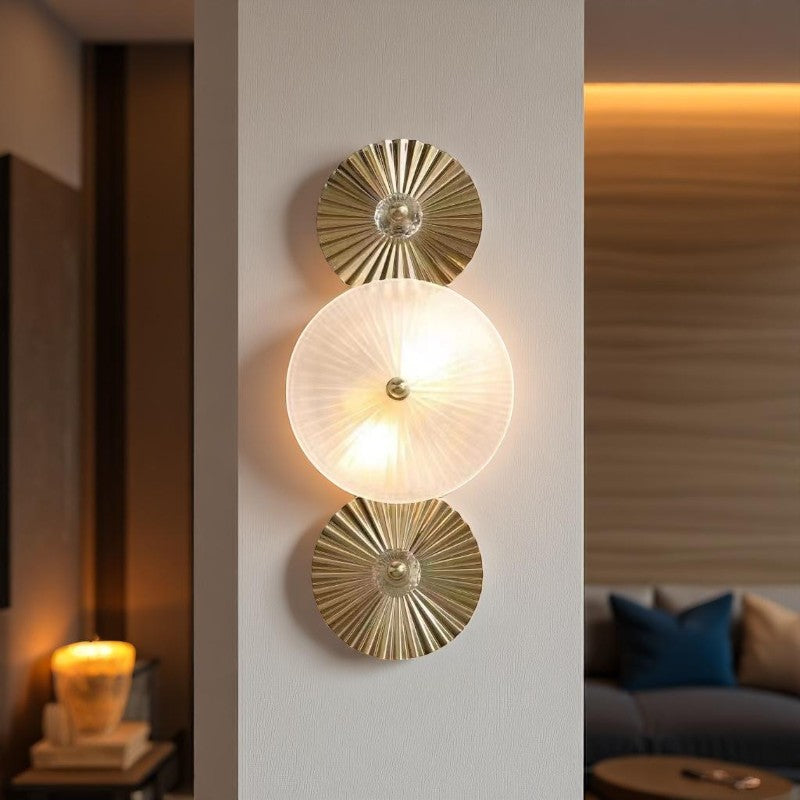 Modern wall sconce featuring a central frosted glass disc that emits a soft, ambient glow, flanked by two decorative, gold-toned metal discs with a sunburst pattern, mounted vertically on a white wall in a contemporary interior setting.