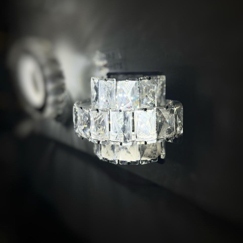 A compact, cube-shaped crystal wall sconce with a sparkling effect, mounted on a dark background.