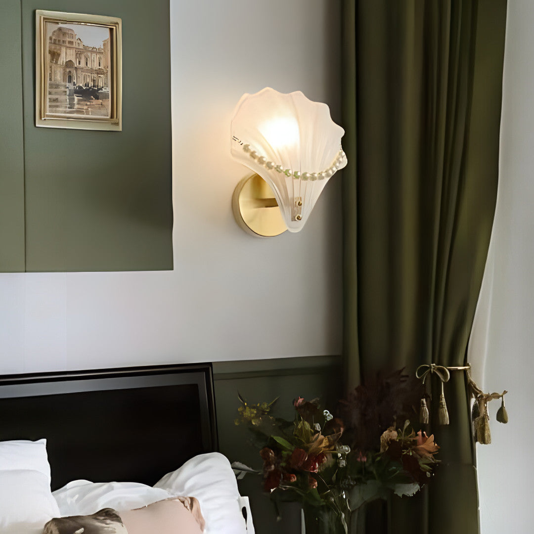 A decorative seashell-shaped wall sconce with a gold circular base and a sculpted white glass shell adorned with pearl-like accents, emitting a warm glow.