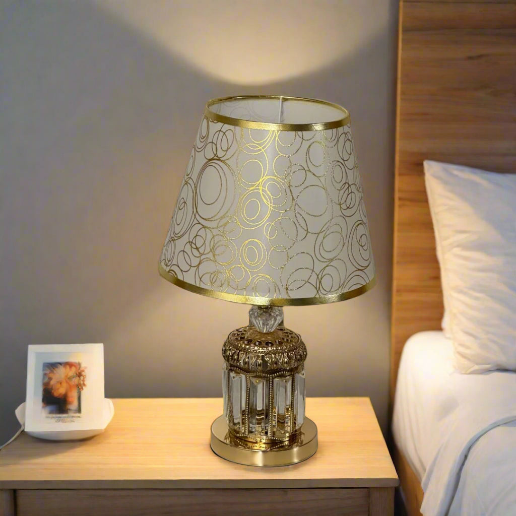 A table lamp with a decorative gold and transparent glass base, and a beige lampshade adorned with gold circular patterns, placed on a wooden surface next to a bed