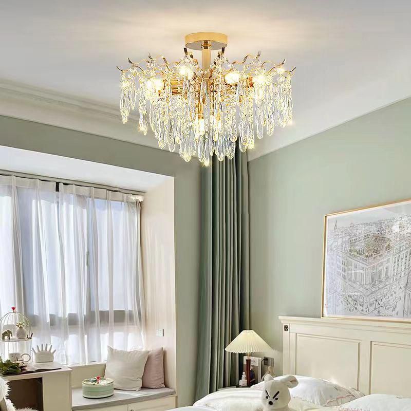 A compact, gold-framed chandelier with cascading crystals installed in a cozy, softly lit bedroom. The chandelier complements the soft green walls, floor-length curtains, and a bed-side art frame.