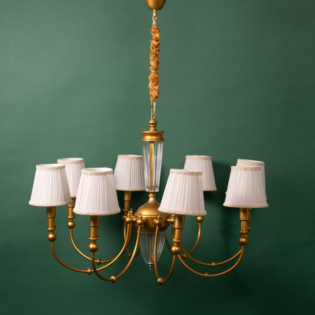A golden chandelier with multiple curved arms, each holding a white pleated lampshade with decorative trim. the chandelier features a central glass column and is suspended from a gold-colored rod.