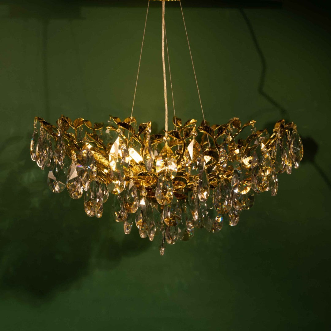 A golden chandelier with a leaf-shaped design and crystal accents, illuminated with a warm yellow glow against a green background.