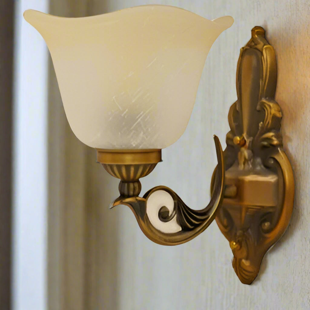 A classic gold wall sconce with a curved arm and frosted glass shade, featuring intricate detailing on the mounting plate.