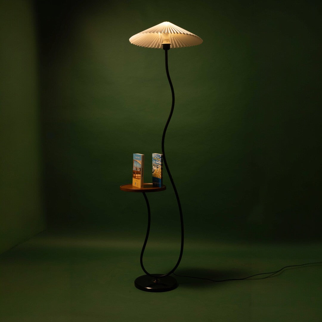 A black floor lamp with a wavy stand, a pleated white lampshade, and a small wooden shelf holding books, illuminated and casting a warm light.