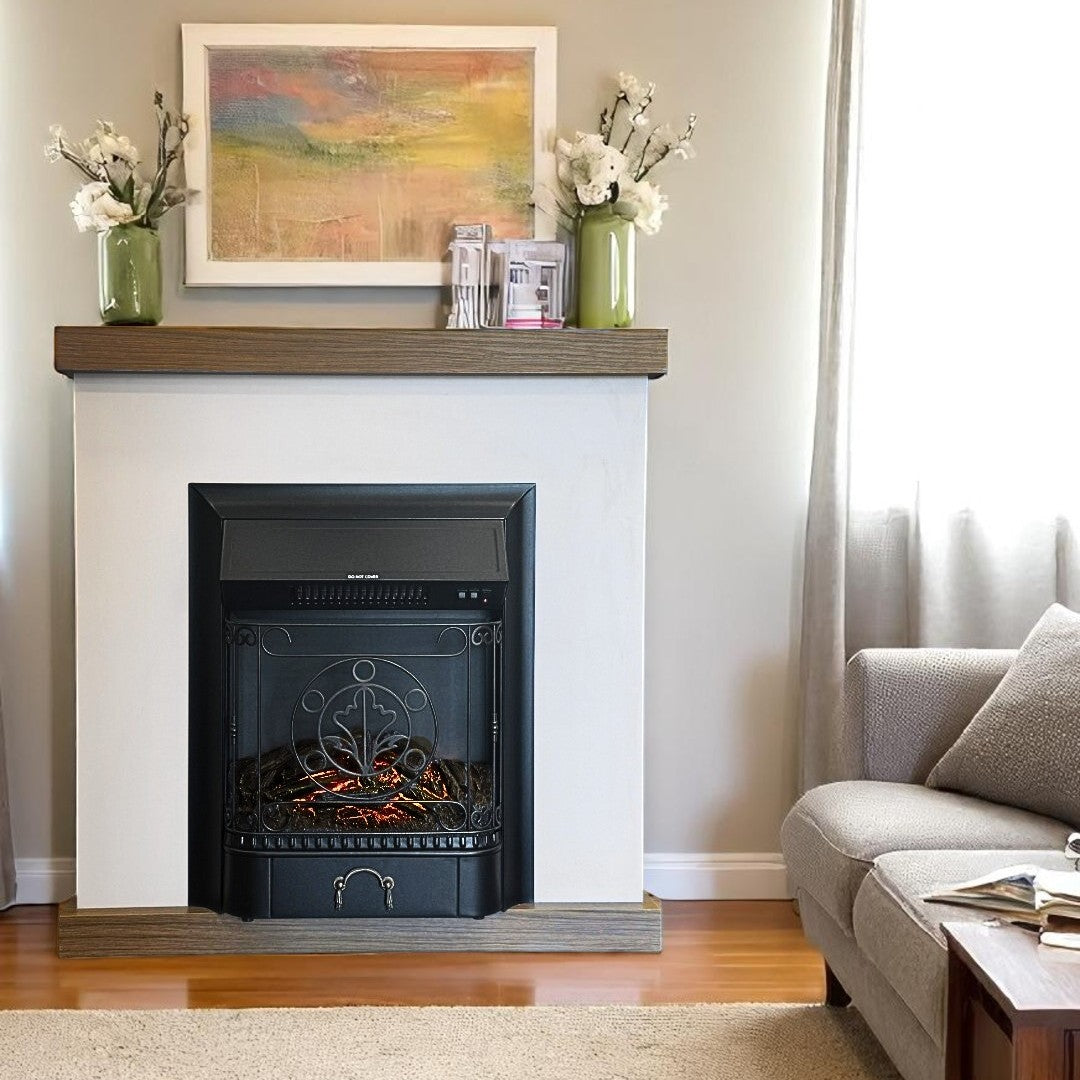 A cozy living room corner featuring a built-in electric fireplace framed with a minimalist wood-trimmed mantel. The fireplace has a black metal front with decorative scrollwork and glowing faux embers. On the mantel, two green vases with white flowers flank a framed abstract painting in warm tones. A soft Gray armchair and a wooden coffee table with magazines complete the inviting setting, bathed in natural light from a nearby window.