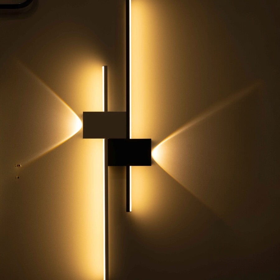 A modern geometric wall sconce with a black and white finish, emitting a dynamic glow with intersecting light beams on a dark background.