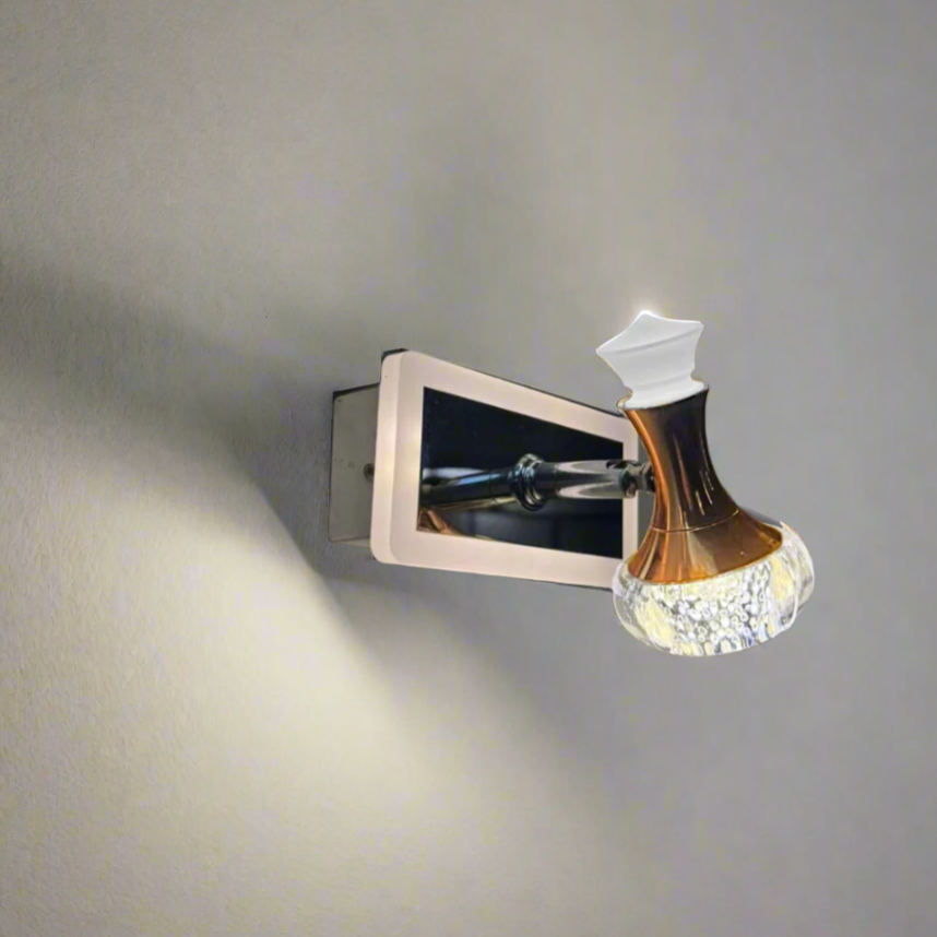 A modern wall-mounted LED light fixture with a metallic rectangular base and an ornate, bottle-shaped glass lampshade that has a golden-brown upper section and a textured, translucent lower section. The fixture is mounted on a plain, light-colored wall and emits a soft, downward glow.