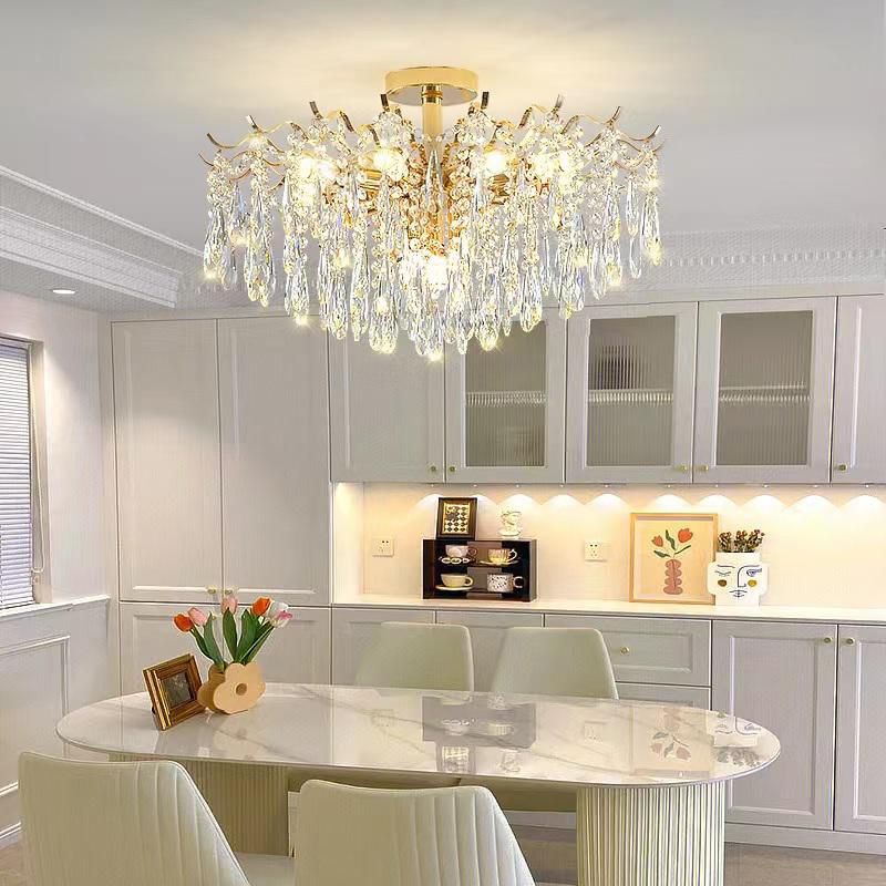 A wide crystal chandelier with gold metal fittings installed above a kitchen dining area. The room features a round white dining table and built-in cabinetry, with warm white lighting enhancing the luxurious yet homey ambiance.