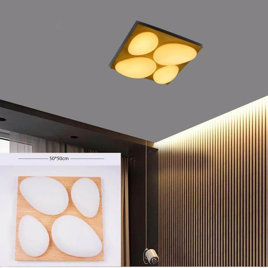 A square ceiling fixture with four rounded triangular lights arranged in a clover shape, set on a beige ceiling corner above vertical wood paneling; it has a golden frame and produces a warm yellow glow