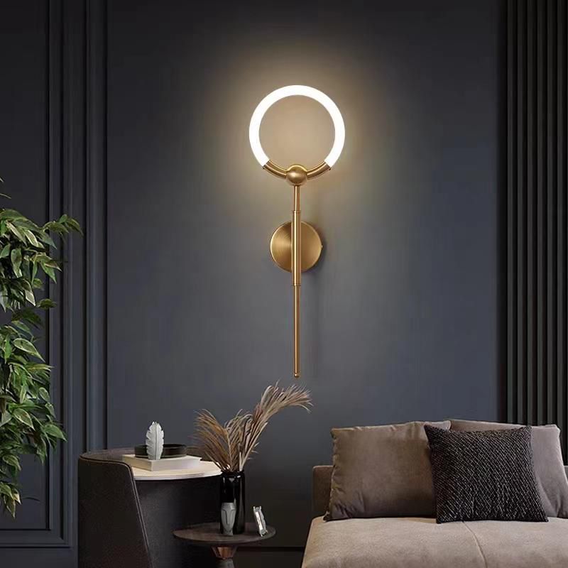 A vertical gold wall lamp featuring a circular LED ring and a slim elongated bar, casting soft ambient light in a dark-themed living space.