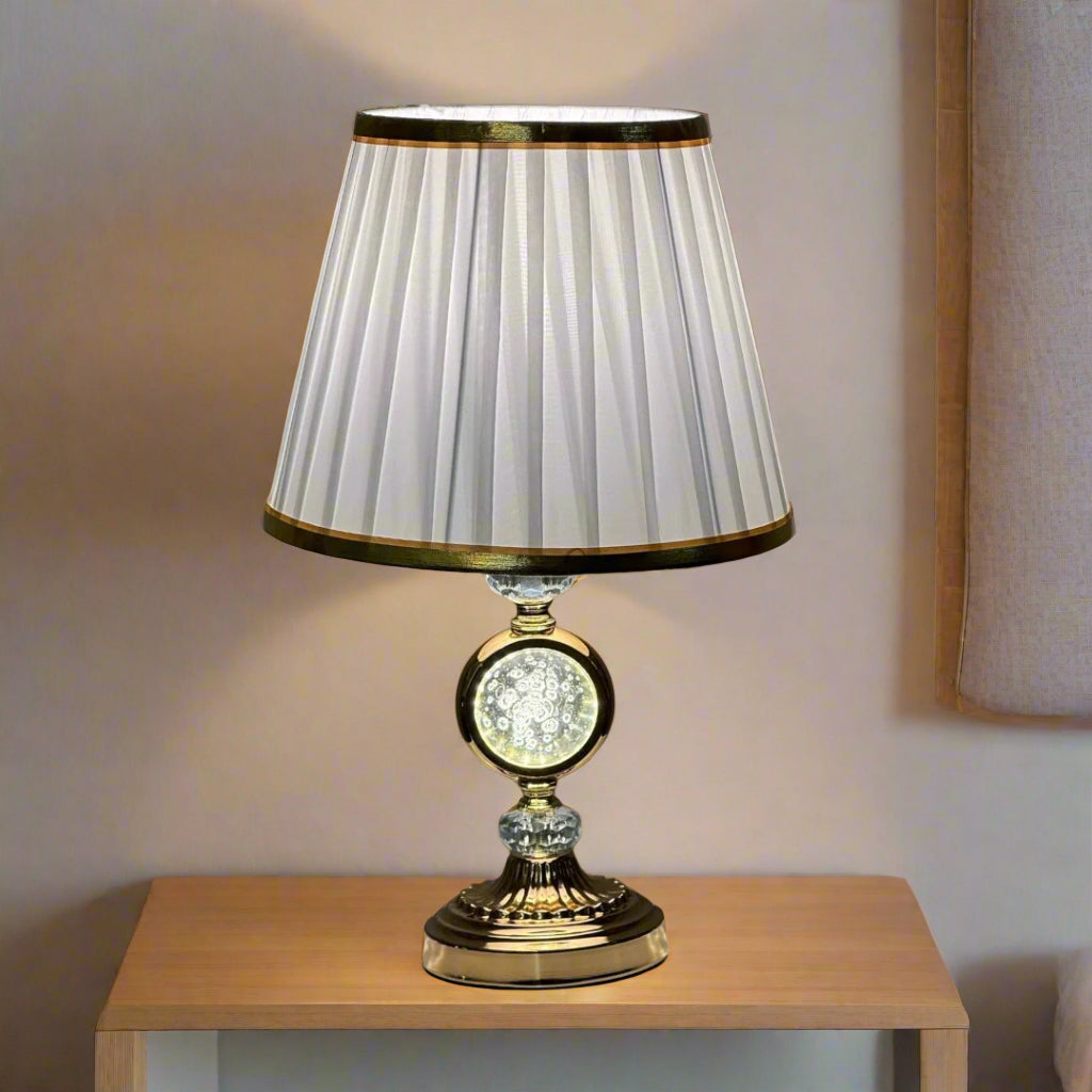 Elegant gold table lamp with a pleated white shade with a gold trim and a crystal accent in the center, placed on a wooden surface against a wood-paneled background.