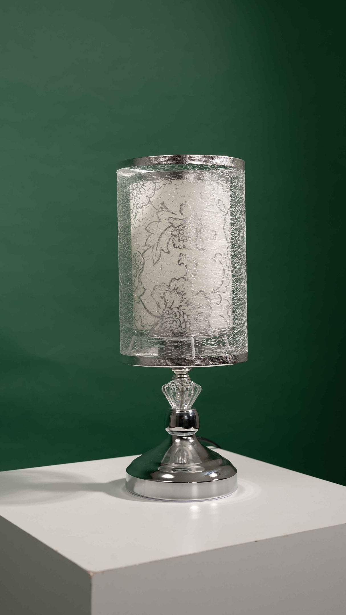 Silver table lamp with a cylindrical transparent shade decorated with a leaf-like etched design, placed on a white table and green background.