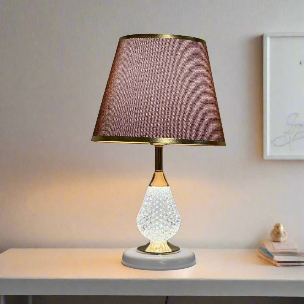 A table lamp with a white textured glass base, gold accents, and a brown fabric lampshade with delicate gold trim, placed on a white desk