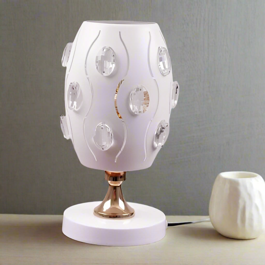 A white globe table lamp with a geometric design, adorned with crystal beads, gold accents, and a sleek base, sits on a wooden table. It features an LED light with an E27 bulb, casting a warm, stylish glow.