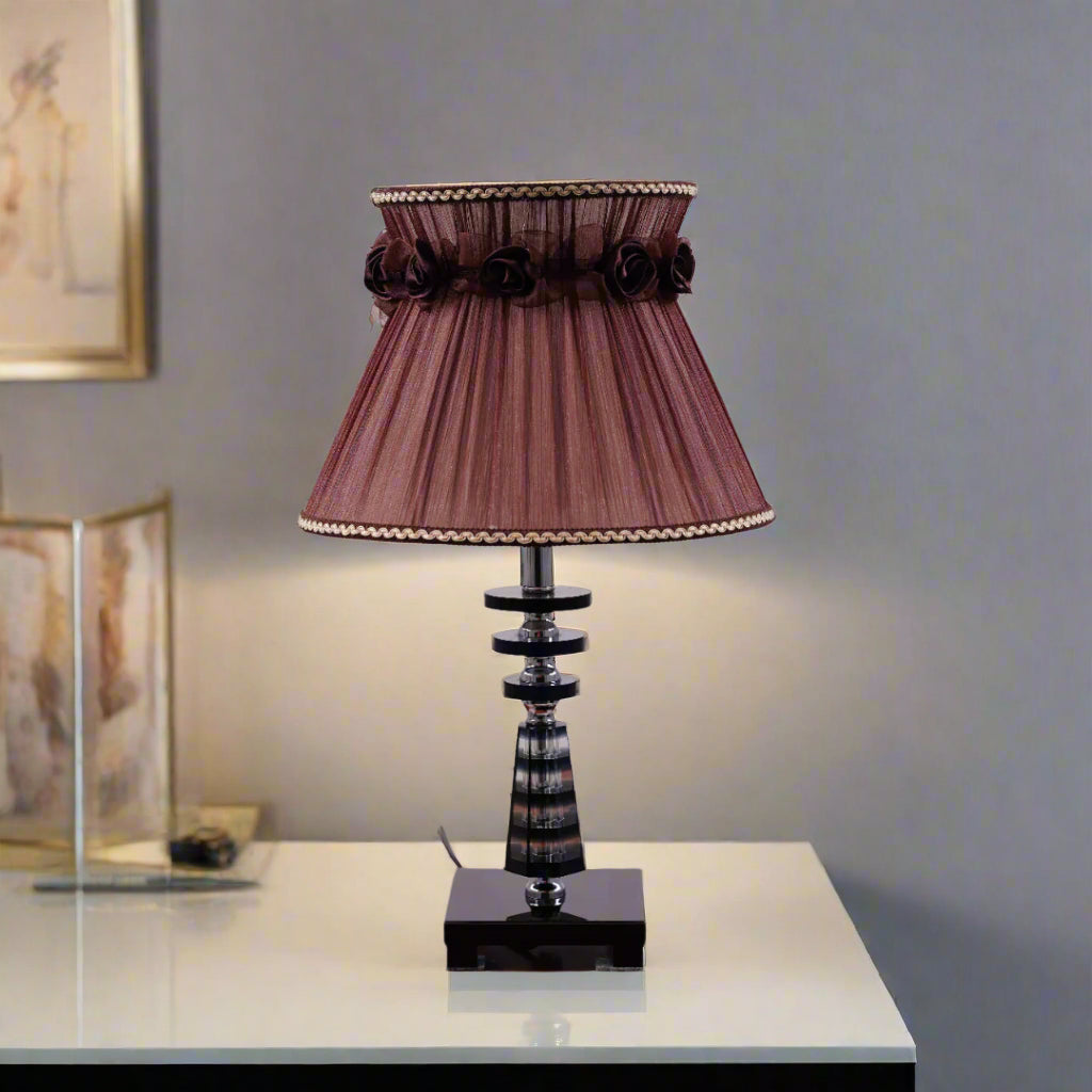 Elegant table lamp with a Brown pleated lampshade, adorned with delicate fabric roses and gold trim. The base is crafted from a combination of transparent and amber-colored glass with a geometric design, supported by a sturdy glass platform. Equipped with an E27 LED light, it emits a warm and sophisticated glow.