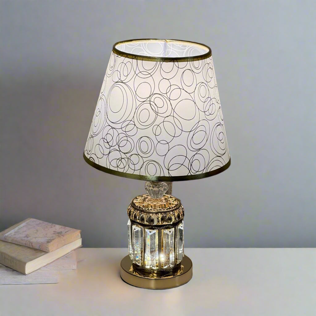 A vintage-style table lamp with a gold and glass base, and a white lampshade featuring a modern circular pattern, placed on a white surface with soft lighting.