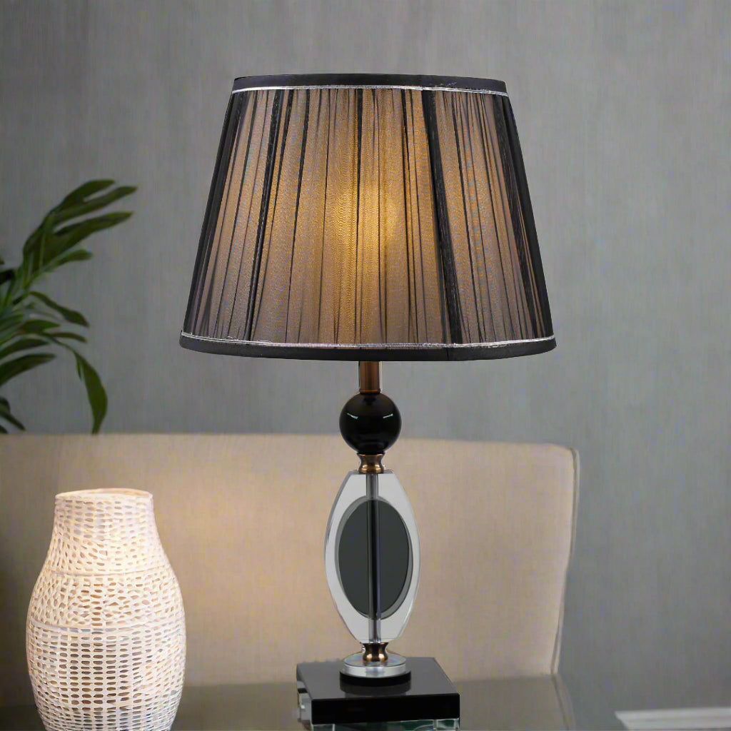 An elegant table lamp with a black pleated lampshade, equipped with an E27 LED light, emits a warm and sophisticated glow. The base is crafted from a combination of gold metal and transparent black glass with a geometric design, supported by a sturdy black glass platform. placed on a glass table.