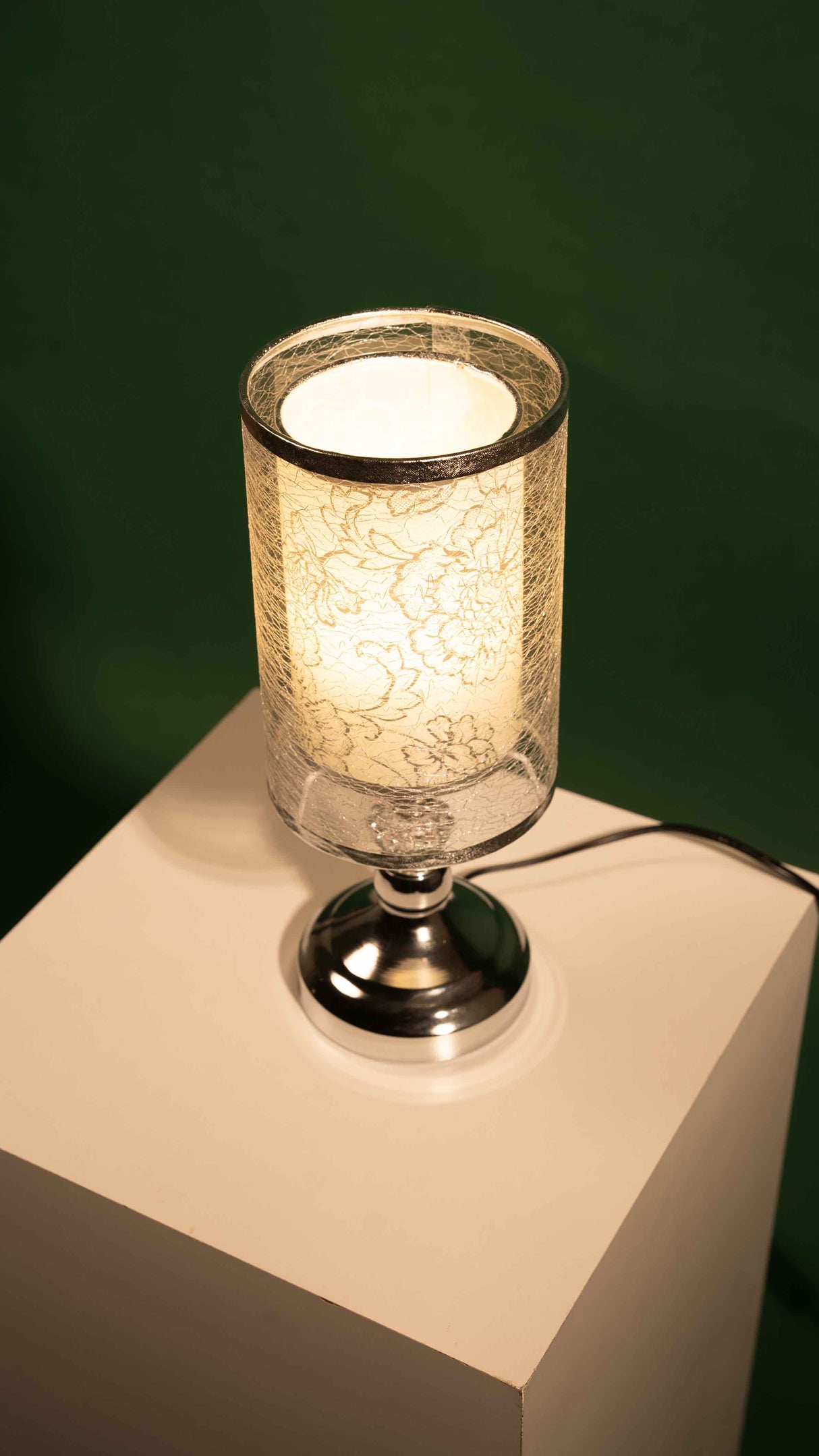 A modern table lamp with a cylindrical shade featuring delicate lace patterns, a silver base, and a warm glowing light equipped with an LED lamp E27, placed on a white surface against a dark green background.