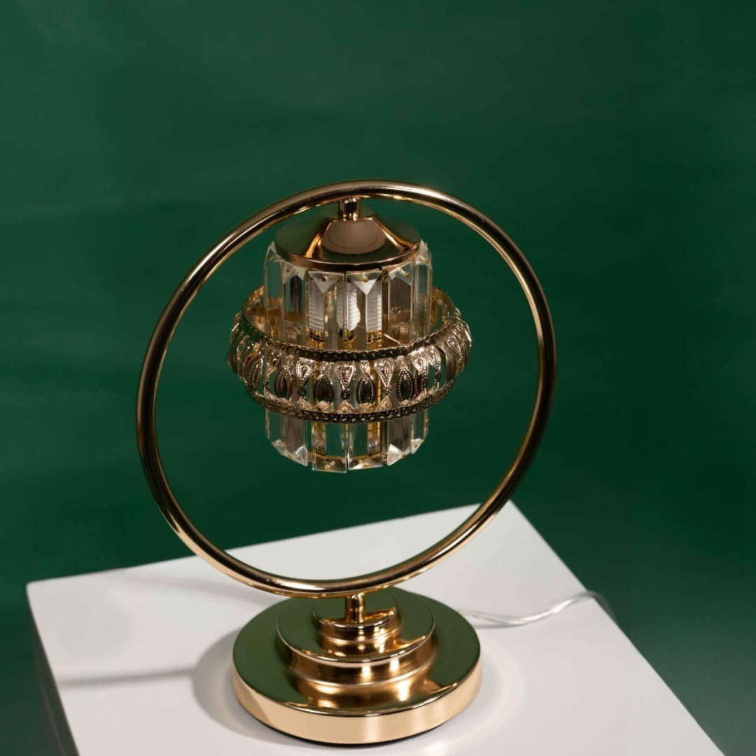 A luxurious table lamp with a circular gold frame and suspended crystal elements, designed to resemble a chandelier. Placed on a white table and green background.