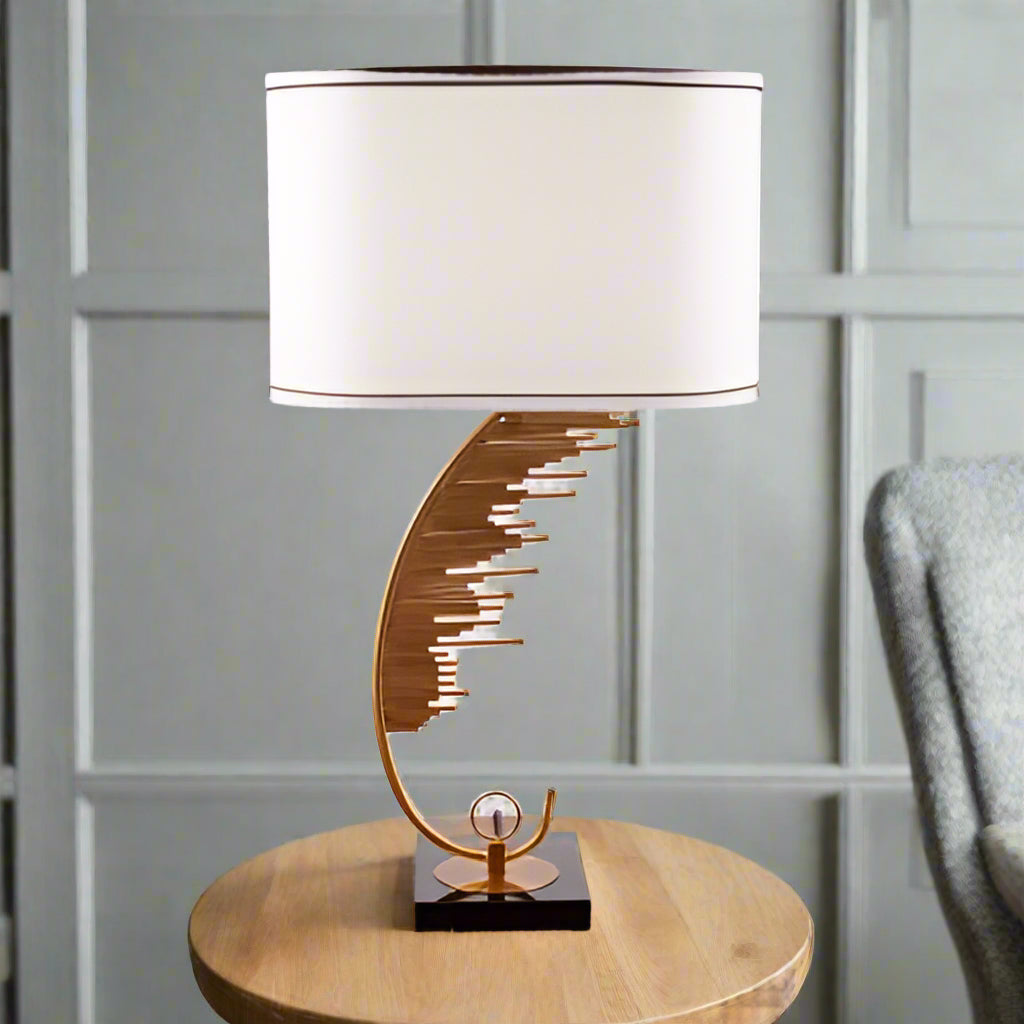 A contemporary table lamp with a gold metal base shaped like a curved staircase or an abstract wave pattern. The base is mounted on a black square platform. The lamp has a large, cylindrical white fabric shade with a thin brown trim at the edges.