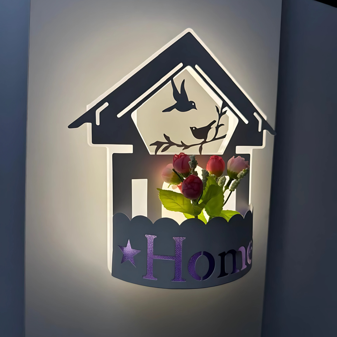 Wall-mounted LED light fixture designed in the shape of a white house , featuring a cork-style built-in compartment holding artificial pink and red tulip flowers. The light has a whimsical, cottage-inspired aesthetic and is mounted on a plain white wall.