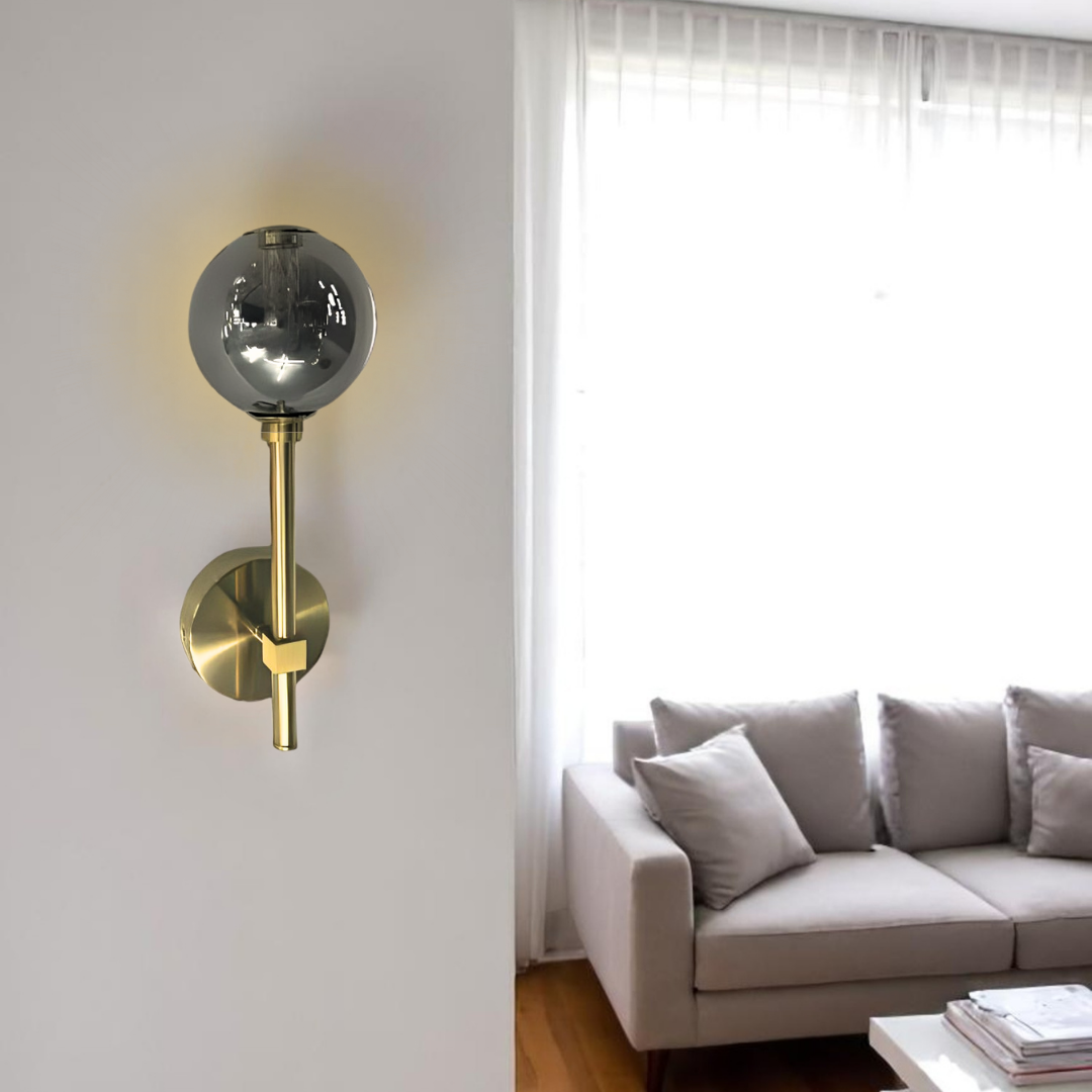A modern wall sconce with a gold metal base and a reflective smoked glass globe shade mounted on a white wall. In the background, a bright living room features a beige sofa with multiple throw pillows, sheer white curtains, and a white coffee table with stacked magazines.