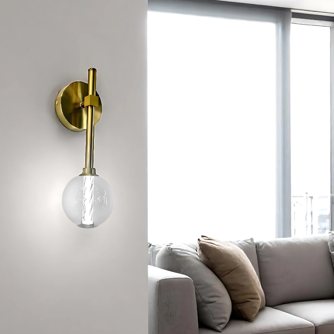 A modern wall sconce with a gold metal base and glass globe shade mounted on a white wall. In the background, a bright living room features a beige sofa with multiple throw pillows, sheer white curtains, and a white coffee table with stacked magazines.