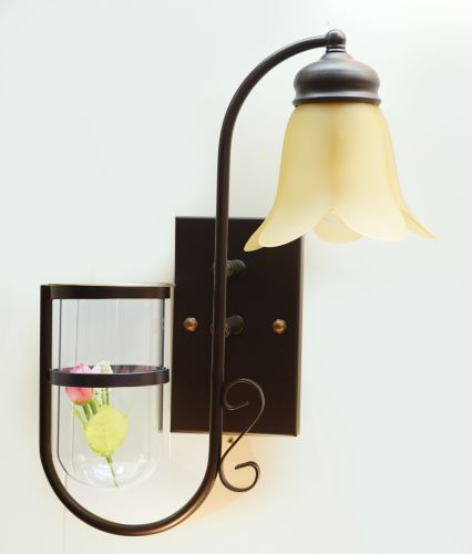A rustic-inspired wall light with a brown metal frame, a curved arm, and a frosted glass shade in a flower-like shape . Below it, a small decorative glass shelf holds a flower.