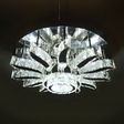 A grand round chandelier with multiple outward-bending arms made of clear crystal, forming a radial pattern and suspended from a dark ceiling; it gives off cool white lighting and adds dramatic flair to large spaces.

