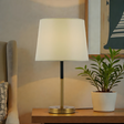 Table lamp with white shade and gold base on a wooden table next to a plant, with a neutral wall and framed picture in the background.
