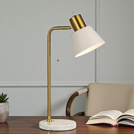 Table lamp with gold base and white shade on a desk with an open book and plant.