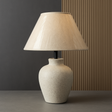 Beige table lamp with a textured base against a dark wall.