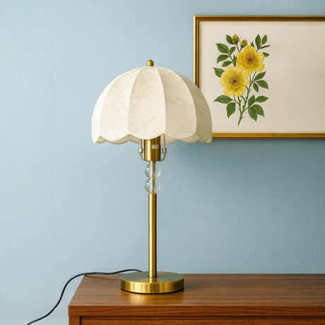 Table lamp with white shade and gold base on a wooden surface, with a floral painting in the background.