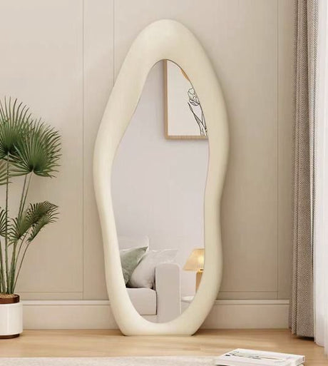 Egg-shaped mirror on a wooden floor with a plant and framed picture in the background.