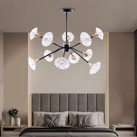 A striking modern chandelier with black metal arms, gold-toned light sockets, and intricate flower-shaped glass shades serves as the focal point of a stylish, minimalist bedroom. The room features a tall, padded Gray headboard, neatly arranged pillows, and neutral-toned walls that complement the chandelier’s elegant design.