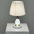 A table lamp with a crystal-textured white metal base, gold accents, and a soft beige fabric lampshade, placed on a white table against a grey wall.