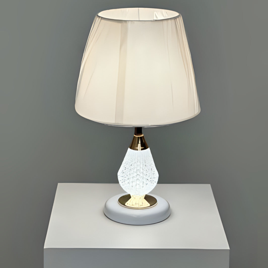 A table lamp with a crystal-textured white metal base, gold accents, and a soft beige fabric lampshade, placed on a white table against a grey wall.