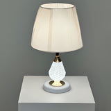 A table lamp with a crystal-textured white metal base, gold accents, and a soft beige fabric lampshade, placed on a white table against a grey wall.