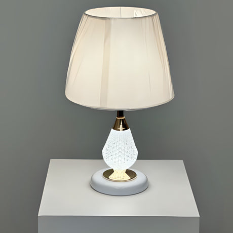 A table lamp with a crystal-textured white metal base, gold accents, and a soft beige fabric lampshade, placed on a white table against a grey wall.