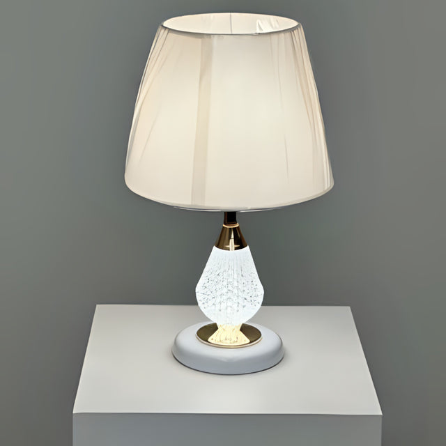 A table lamp with a crystal-textured white metal base, gold accents, and a soft beige fabric lampshade, placed on a white table against a grey wall.