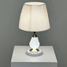 A table lamp with a crystal-textured white metal base, gold accents, and a soft beige fabric lampshade, placed on a white table against a grey wall.