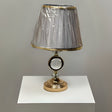 Round gold table lamp with a pleated grey shade with gold trim, featuring a crystal centrepiece, placed on a white table.