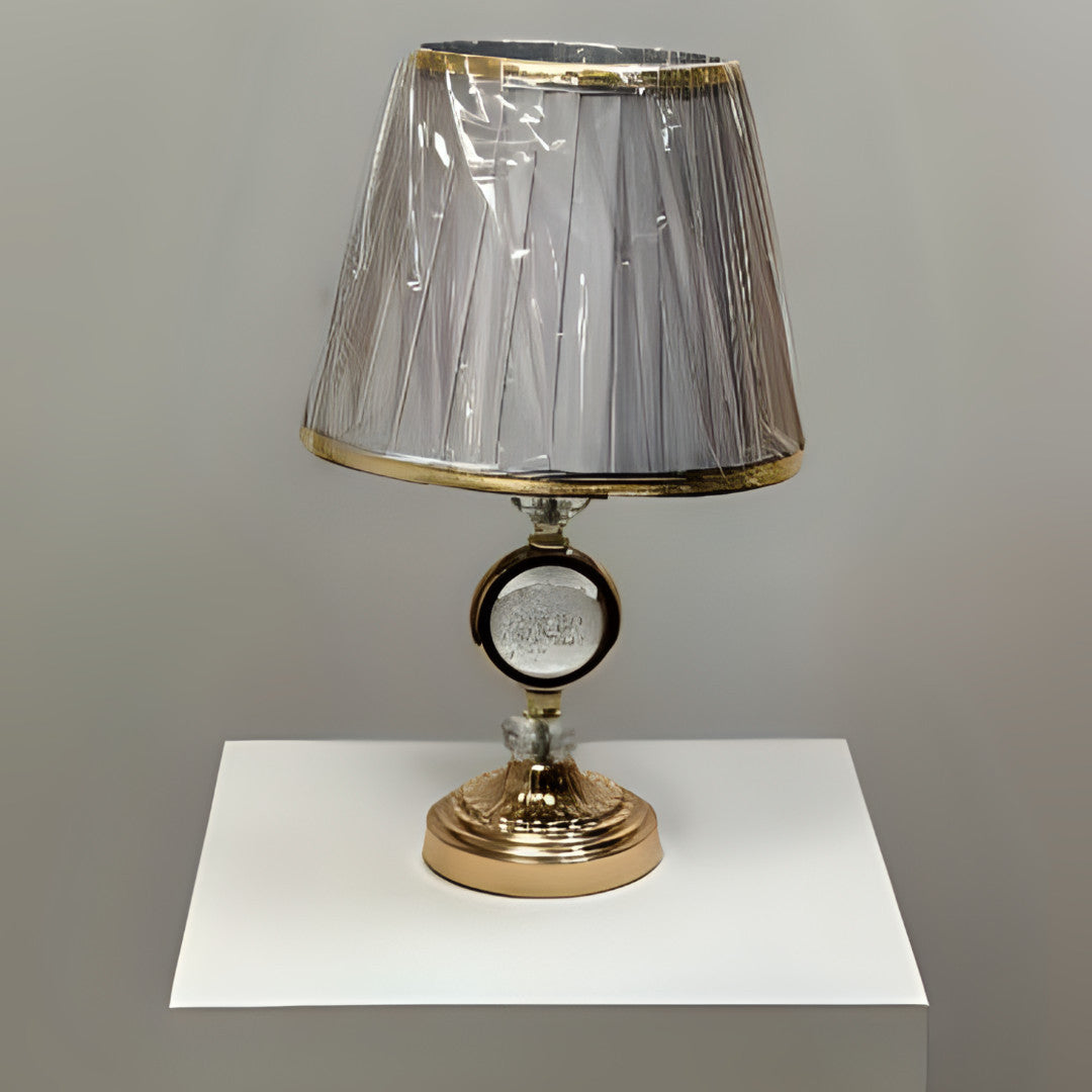 Round gold table lamp with a pleated grey shade with gold trim, featuring a crystal centrepiece, placed on a white table.