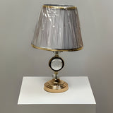 Round gold table lamp with a pleated grey shade with gold trim, featuring a crystal centrepiece, placed on a white table.