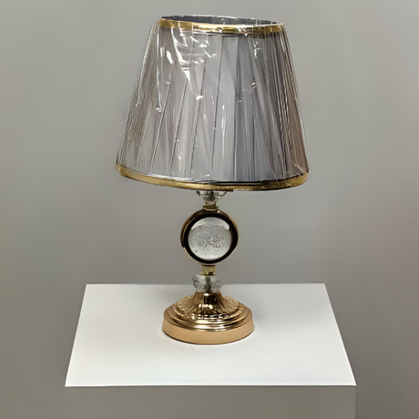Round gold table lamp with a pleated grey shade with gold trim, featuring a crystal centrepiece, placed on a white table.