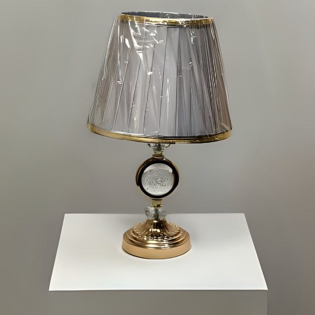 Round gold table lamp with a pleated grey shade with gold trim, featuring a crystal centrepiece, placed on a white table.